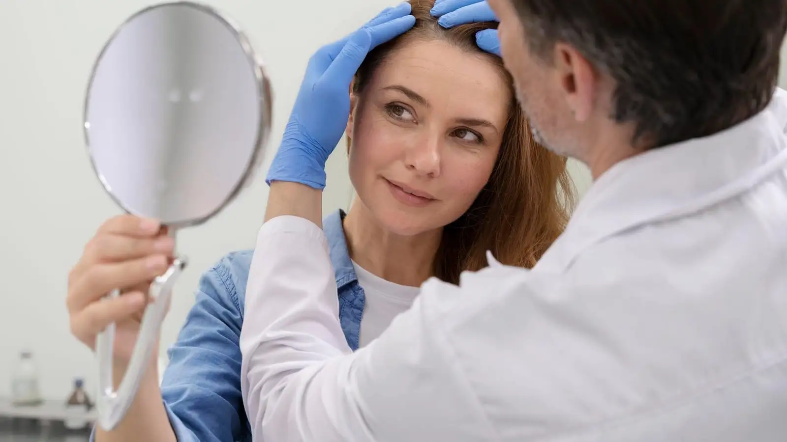 A woman with long hair examining her parting and hairline in a mirror during a hair loss consultation